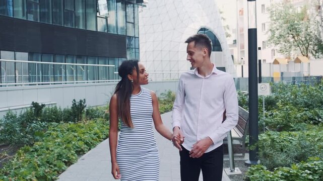 A multiracial couple embraces and kisses while walking along a paved path in a modern urban outdoor setting with greenery and contemporary architecture.