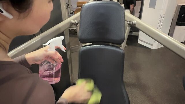 Centered frontal view footage of an Asian woman wiping the padded seat and backrest of an exercise machine using cleaning spray and a microfiber cloth in a modern fitness center.