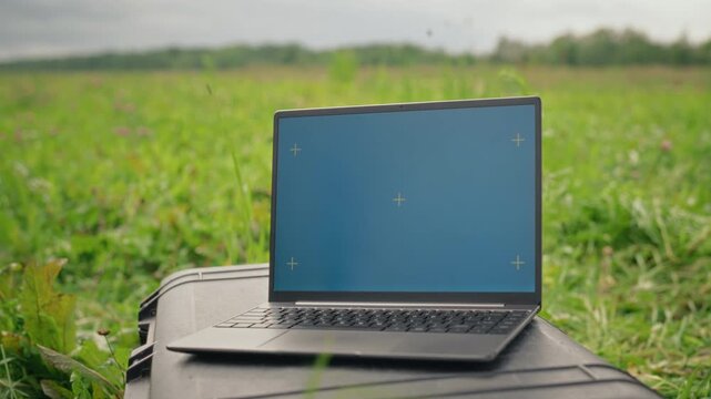 Laptop featuring blue chroma key screen with tracking markers standing outdoors in green field. Footage suits agritech promotion, freelance workflow, and digital software mockup integration