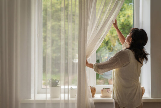Woman standing by a window, opening sheer curtains and welcoming morning sunlight into her domestic interior space