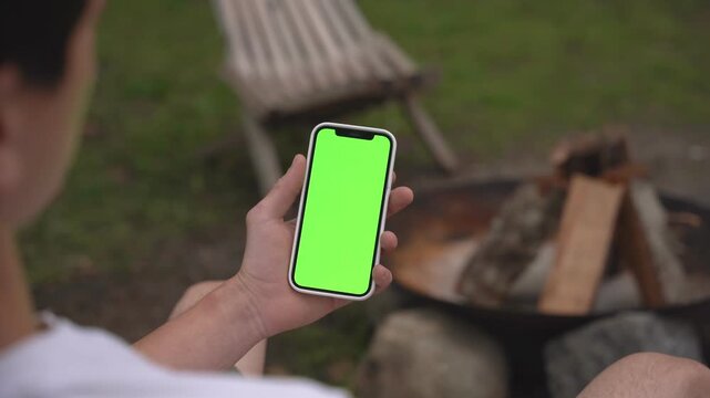 Person holds phone with green screen. Seated near fire pit outdoors. Wood stacked, chair behind. Calm, relaxed mood. Ideal for apps, travel, or social media ads