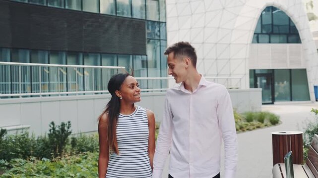 A man and woman holding hands and walking together in a modern urban outdoor plaza during daytime. They look at each other affectionately amid contemporary architecture and greenery.