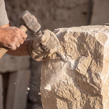Stone mason carving a rough rock block with a metal chisel and hammer mallet, generative AI