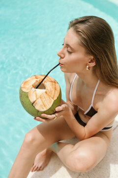Woman drinking fresh coconut beside a tropical swimming pool