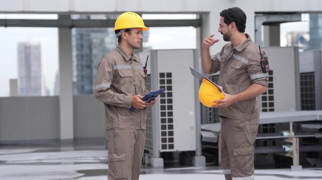 Technician worker wearing hard hat uses tablet for rooftop air conditioner inspection and maintenance, coordinating equipment service beside outdoor cooling unit on modern building roof
