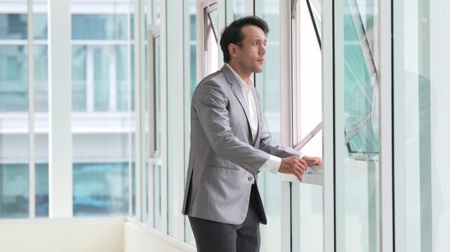 Businessman in gray suit beside office window, thoughtful professional portrait of man in modern corporate building architecture, walking through bright workplace corridor with confident urban outlook