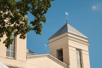 Beige church bell tower with tall arched windows, gray hipped roof and metal cross, with a second tower and roofline partly visible among green oak branches against a clear blue sky © Vasilisa Dorofeeva