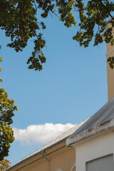 Church roof with a sloped light roof surface, metal gutter and beige cornice framed by dense green oak and maple branches against a deep blue summer sky with a fluffy white cloud © Vasilisa Dorofeeva