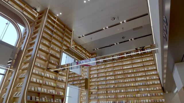 A modern multi-level indoor library with extensive wooden bookshelves filled with books, featuring ceiling lighting fixtures.