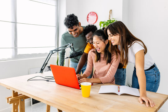 Group of diverse college friends gathered around a laptop and microphone recording a podcast at home. Young multiracial people laughing together. Friendship and technology concept.