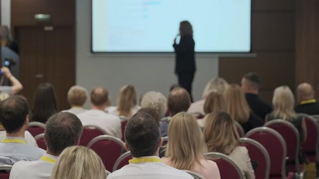 Audience listening to presenter during business conference, people seated in rows watching presentation on screen.