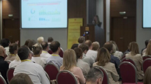 Business audience seated in rows, listening to a presenter at a corporate conference, slides visible on a projection screen, professional training and networking atmosphere.