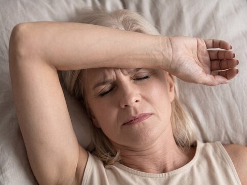 Mature woman suffering from a severe migraine headache lying in bed with her arm over her forehead.