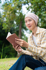 Asian female cancer survivor with earphones holding a book and looking at nature in green park. Finding peace, mental wellbeing, hopeful recovery, and relaxed lifestyle concept. Vertical Shot