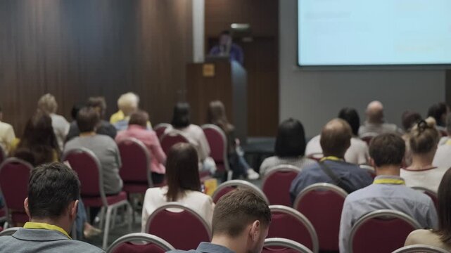 Conference audience listening to presenter, professionals taking notes and watching projected slides, corporate seminar atmosphere, networking and learning.