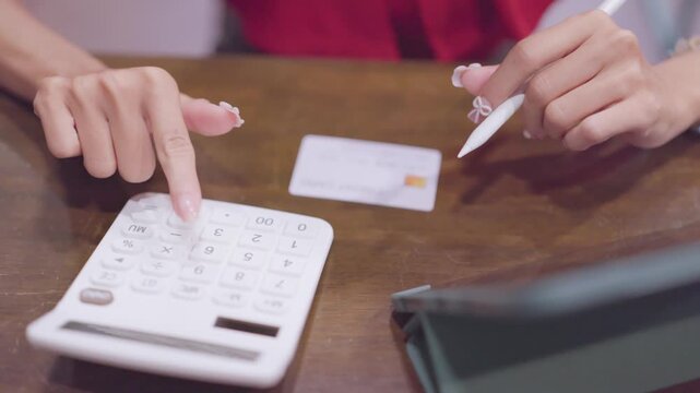 Woman calculates home expenses pressing buttons on calculator holding digital stylus pen next credit card on wooden table indoors.