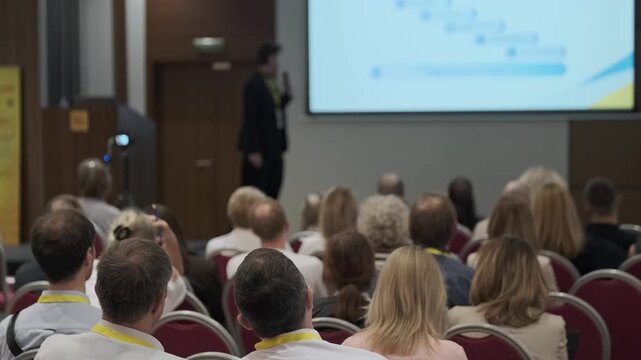 Audience listening to business presentation in conference room, speaker on stage with projection screen, professional seminar atmosphere.