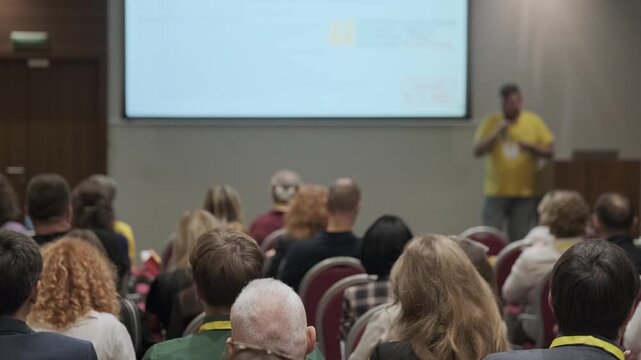 Audience listening to presenter during business seminar, professionals seated in rows, focus on learning, networking, professional development.