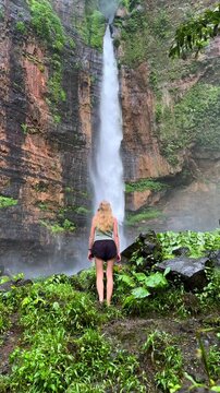 Young woman exploring Kapas Biru Waterfall in East Java, Indonesia, surrounded by lush tropical vegetation and towering rocky cliffs. Scenic travel, adventure and nature destination.