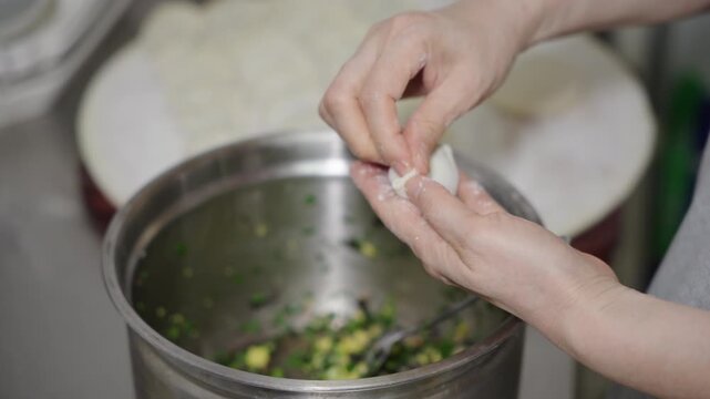 A person's hands are folding a dumpling filled with vegetable filling, over a metal bowl of more filling.