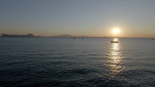 A serene seascape at sunset, with a small boat on calm water and a distant coastal skyline under a gradient sky.
