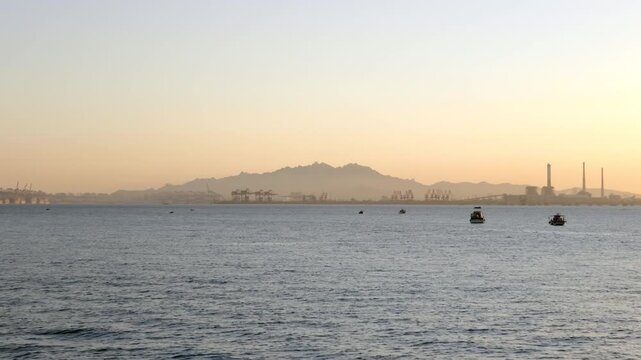 Calm coastal waters at dawn, with small boats and distant mountain silhouettes under a hazy sky.