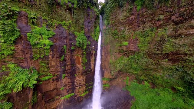 Dynamic FPV drone flight over Kapas Biru Waterfall and along a jungle stream in East Java, Indonesia, revealing a tall cascade, rocky cliffs and lush tropical rainforest.