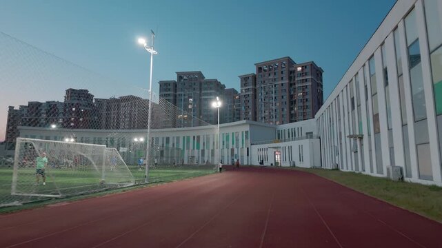 A sports field with running track, soccer goal, and surrounding buildings, captured at dusk with bright floodlights.