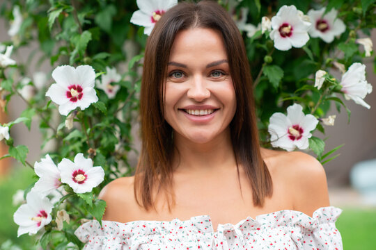 Smiling young woman in a floral off-shoulder dress surrounded by blooming white hibiscus flowers in a garden. Natural beauty portrait with a cheerful summer atmosphere
