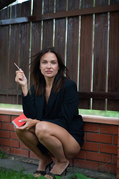 Elegant woman in a black blazer and heels sitting outdoors, holding a red book and cigarette against a rustic wooden fence. Fashion portrait with a sophisticated, contemplative mood