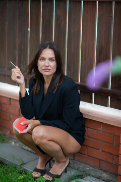 Elegant woman in a black blazer and heels sitting outdoors, holding a red book and cigarette against a rustic wooden fence. Fashion portrait with a sophisticated, contemplative mood