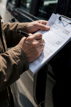 Delivery worker signs clipboard by car. Person confirms package receipt using pen on document. Professional courier confirms delivery, completes paperwork for transit.