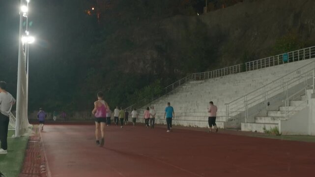 People walk on a red athletic track in an outdoor stadium at night, with bleachers and lighting nearby.