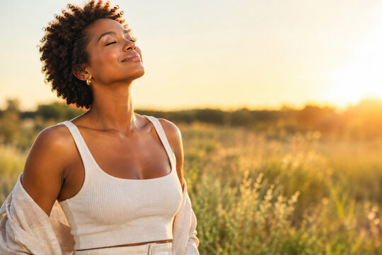 A beautiful young Black woman with curly hair is captured outdoors, her eyes closed in peaceful contentment as she basks in the warm glow of the golden hour sunset