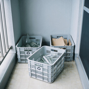 Home balcony recycling corner with three light gray sorting baskets of glass bottles and cardboard boxes
