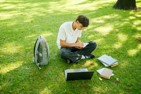 Thoughtful male student writing school tasks while resting in the sunny park.