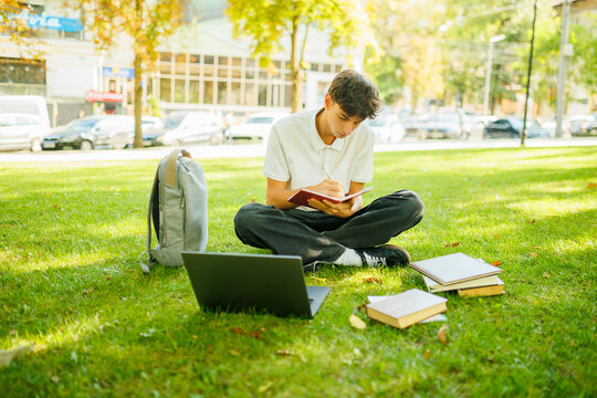 Focused young scholar writing in a notebook while sitting on the campus grass.