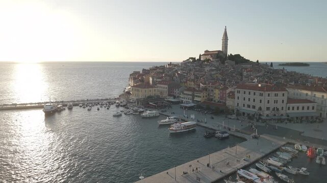 Elevated aerial drone shot showing boats moored along the main harbor pier, lively waterfront square, and historic peninsula rising to St Euphemia bell tower in warm evening sunset light in Rovinj.