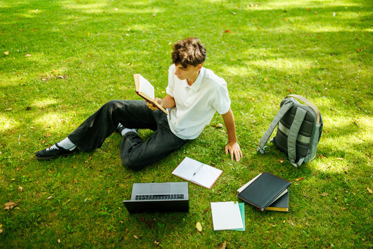 Focused caucasian teenager reading a book while sitting on the campus grass.