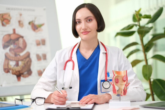 Confident physician working at desk in bright medical office, wearing white coat and blue scrubs, with stethoscope, anatomy model rectum, charts, glasses, and plant in soft daylight during