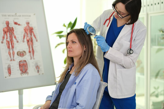 Female doctor wearing gloves and white coat checks woman scalp with handheld magnifier during calm consultation in bright clinic room, suggesting hair care assessment, dermatology, and personal