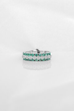 Luxury silver-tone band ring decorated with alternating green and clear sparkling stones, displayed on a reflective white surface.