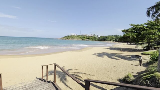 View of a sandy beach with wooden stairs leading down to the shore, turquoise ocean waves, and a couple walking along the coastline in Guarapari, Espirito Santo, Brazil.