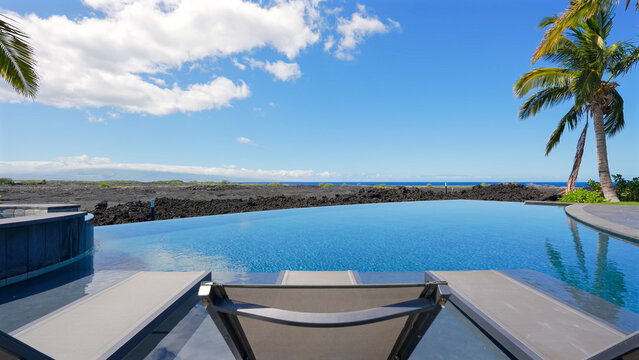 Luxury infinity pool overlooking a dark volcanic rock shoreline, blue ocean, and palm trees under blue sky
