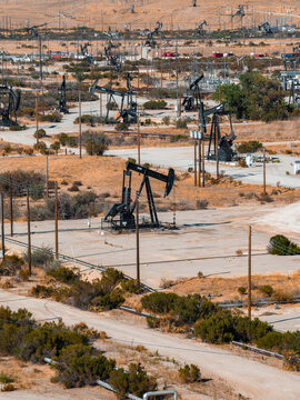 Aerial view shows numerous pump jacks extracting petroleum in a dry California oil field. Infrastructure pipelines and power lines crisscross the arid industrial terrain under daylight.