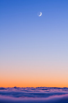 Crescent moon shining in a vibrant twilight sky above a thick layer of soft rolling clouds