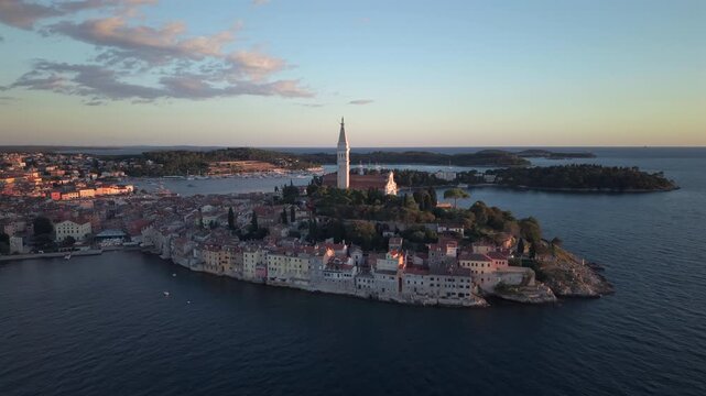 High wide-angle drone shot capturing the historic peninsula of Rovinj glowing in golden evening sunlight, featuring the Church of St Euphemia bell tower and coastal stone houses.