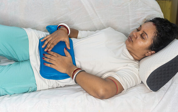Indian woman suffering from heavy menstrual cramps or periods pain, applying a blue hot water bag on her stomach to relieve the pain and feel better, lying on the living room sofa.