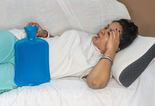 Indian young sick woman checking fever with thermometer during menstrual periods. Using hot water bag to relive cramp or pain, while lying on sofa at home. Fever and illness concept.