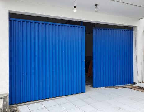 Bright blue corrugated metal double garage doors set in a white wall with a hanging light bulb and white tiled driveway.

